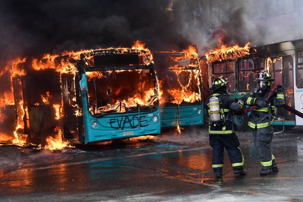 TOPSHOT - Chilean firefighters extinguish a burning bus during clashes between protesters and the riot police in Santiago, on October 19, 2019. - Chile's president declared a state of emergency in Santiago Friday night and gave the military responsibility for security after a day of violent protests over an increase in the price of metro tickets. (Photo by Martin BERNETTI / AFP) (Photo by MARTIN BERNETTI/AFP via Getty Images)