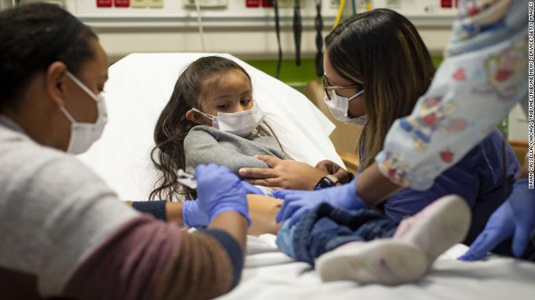 La participante en el ensayo Elena Rosales (al centro), de 3 años, recibe la vacuna Moderna contra covid-19 de manos de la enfermera Lela Lartey (izquierda) con su madre, Mariaelena Lozano (derecha), a su lado, el 7 de diciembre de 2021, en el Hospital Infantil Lurie de Chicago.