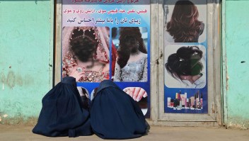 Mujeres afganas vestidas con burka sentadas frente a un salón de belleza con imágenes de mujeres desfiguradas con pintura en aerosol en Jalalabad el 13 de diciembre de 2021. (Wakil Kohsar/AFP/Getty Images)