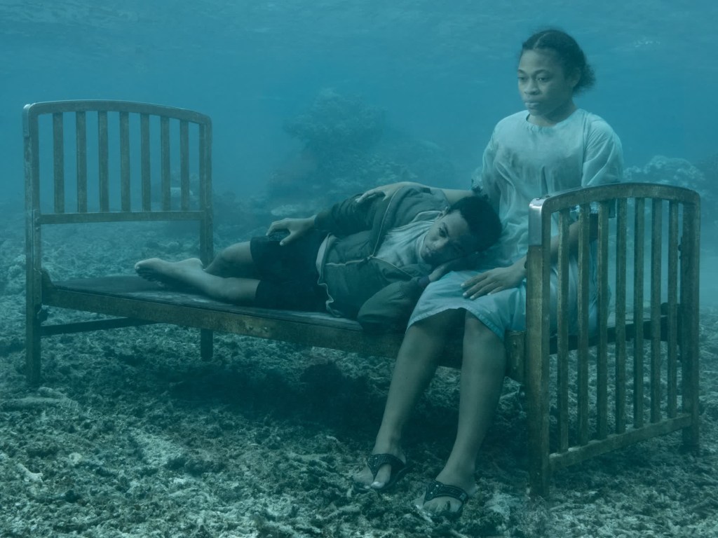 Serafina y Keanan, hermanos, posan uno junto al otro en una cama bajo el agua frente a la costa de Fiyi en la serie "SINK / RISE" del fotógrafo Nick Brandt.