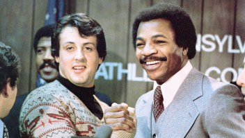Sylvester Stallone y Carl Weathers se dan la mano y sonríen juntos durante una conferencia de prensa en una imagen fija de la película 'Rocky', dirigida por John G. Avildsen, 1976. (United Artists/Getty Images)