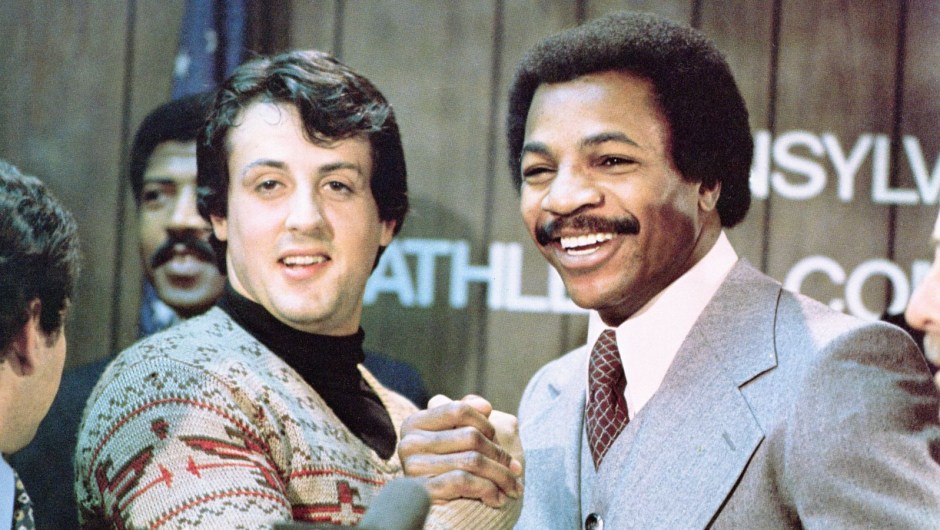 Sylvester Stallone y Carl Weathers se dan la mano y sonríen juntos durante una conferencia de prensa en una imagen fija de la película 'Rocky', dirigida por John G. Avildsen, 1976. (United Artists/Getty Images)