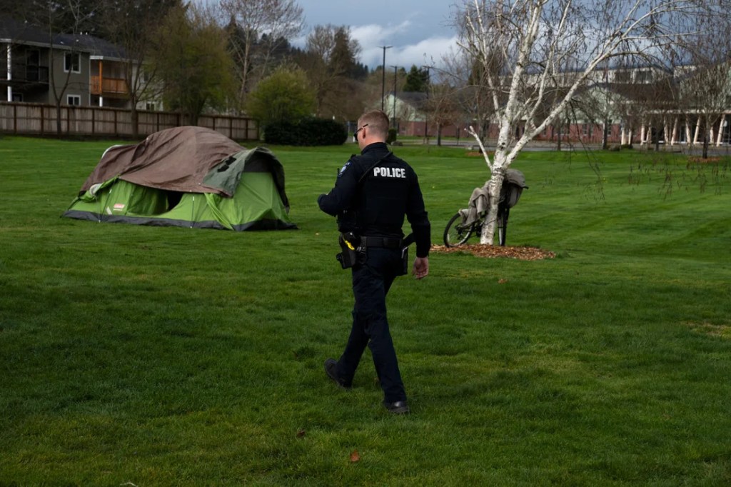 Un oficial de policía de Grants Pass investiga a una persona sin hogar después de que sus familiares solicitaron un control de asistencia social en Fruitdale Park el 23 de marzo de 2024. (Foto: Jenny Kane/AP/Archivo).